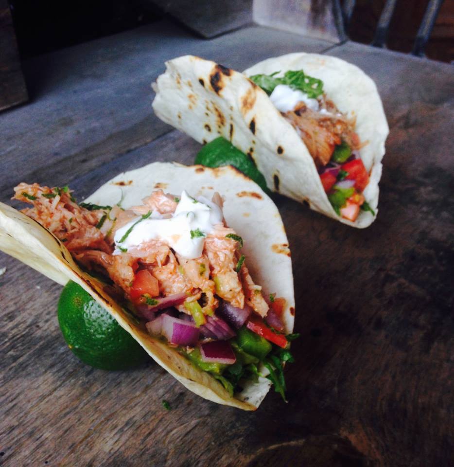 two tacos with chicken and garnish held up with limes on a wooden table