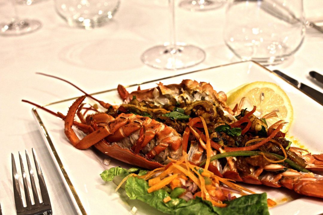 white square plate with full lobster with lemon garnish and wine glasses in the back