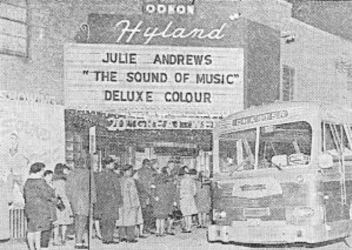 old photo of cinema in black and white with people lined up on sidewalk outside behind bus