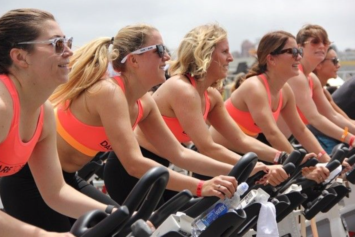 5 women riding stationary bikes outside wearing matching workout outfits